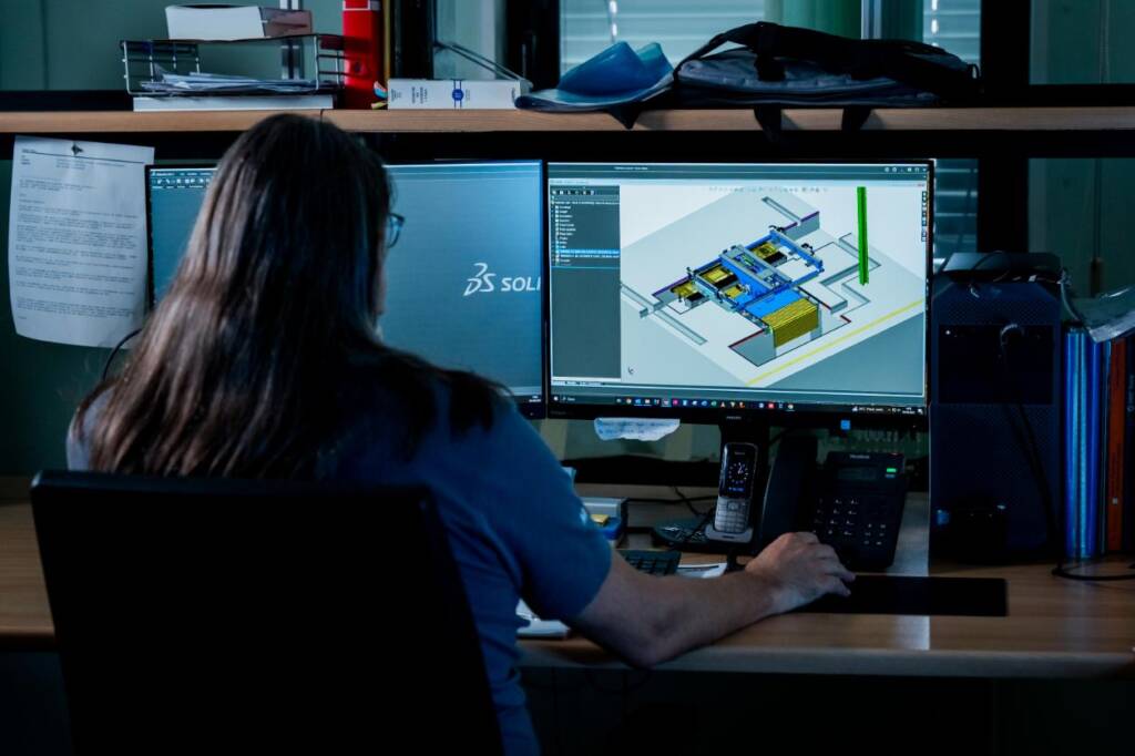 L'immagine mostra un operatore che interagisce con un software di progettazione CAD su un computer, utilizzato per sviluppare soluzioni di automazione industriale. Sullo schermo è visibile un modello tridimensionale di un impianto di produzione, evidenziando elementi come nastri trasportatori e attuatori. L'ambiente di lavoro è attrezzato con strumenti di comunicazione e documentazione, suggerendo un contesto tecnico orientato alla progettazione di