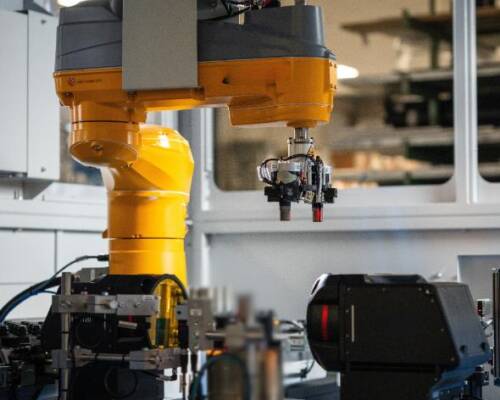 L'immagine mostra una cella robotizzata in un impianto industriale, dotata di un braccio robotico giallo attrezzato con un sistema di presa per la movimentazione di componenti. Sono visibili anche sensori e unità di collaudo, indicativi di un'applicazione nel settore del manufacturing. La configurazione è progettata per ottimizzare l'automazione dei processi produttivi.